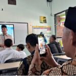 A man in a beige uniform speaks at the front of a classroom or meeting room addressing students seated at desks while a person in the foreground records with a smartphone
